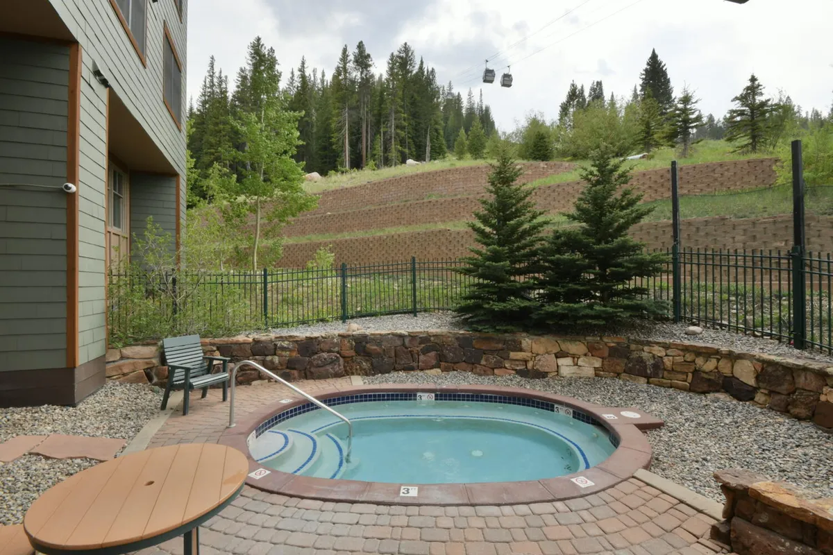An inviting exterior hot tub surrounded by trees and mountain views, with patio furniture nearby. The atmosphere is tranquil, suggesting relaxation, socializing, and soaking in the warmth while gazing at the scenery or watching the gondolas pass overhead.