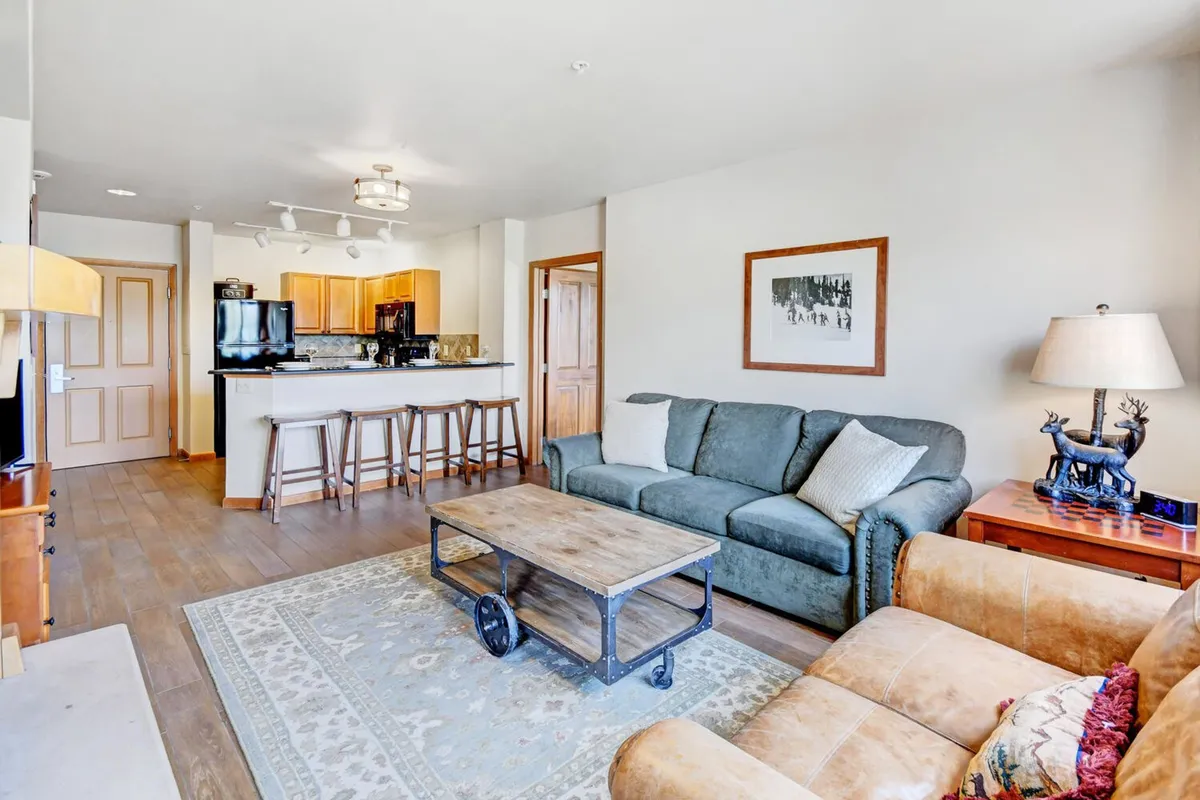 Another angle of the open-concept living space, focusing on the comfortable sofa and coffee table, with the kitchen and dining table visible in the background. Mountain-inspired artwork adorns the walls, and large windows bathe the space in natural light, enhancing the inviting and communal feel.
