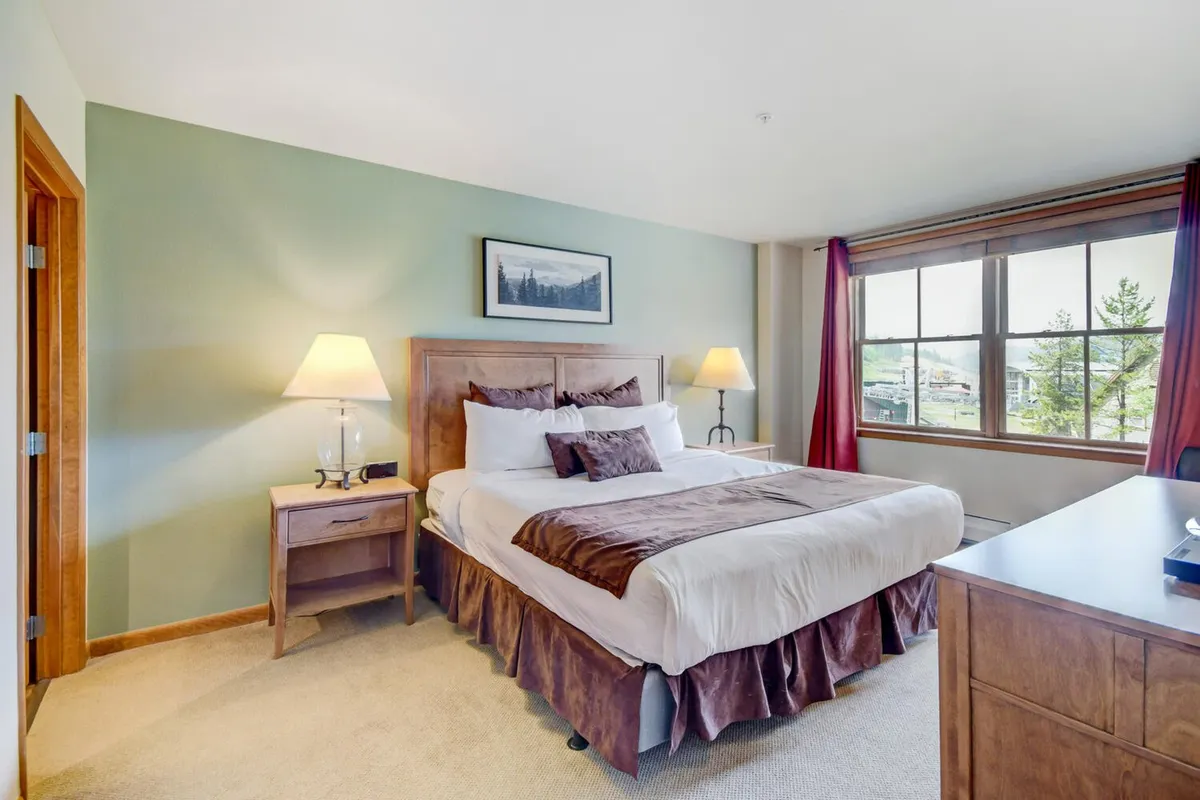 A thoughtfully designed bedroom space showcasing a comfortable bed, window with mountain views, a nightstand, lamp, and art. The soft natural light enhances the room's restful quality, emphasizing comfort and relaxation.