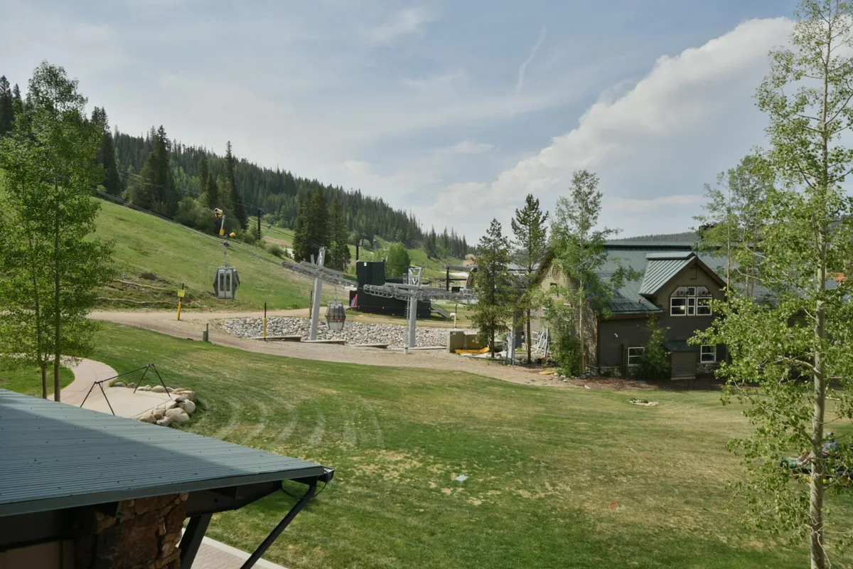 A picturesque exterior view featuring a ski lift against a backdrop of open green space, lush trees, and majestic mountains. The scene is filled with energy, capturing both winter and summer possibilities, from skiing and snowboarding to hiking and picnicking.