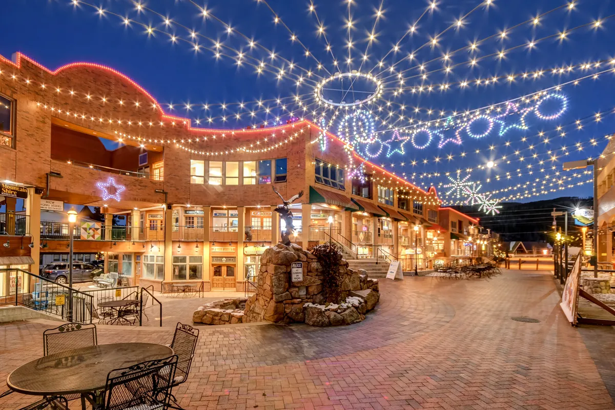A lively exterior scene with overhead string lights illuminating a cobblestone path, decorative stars adding charm, a stone fountain as a focal point, and people subtly engaged in shopping, dining, and socializing. The ambiance suggests the heart of a vibrant mountain village, possibly during a festive evening.