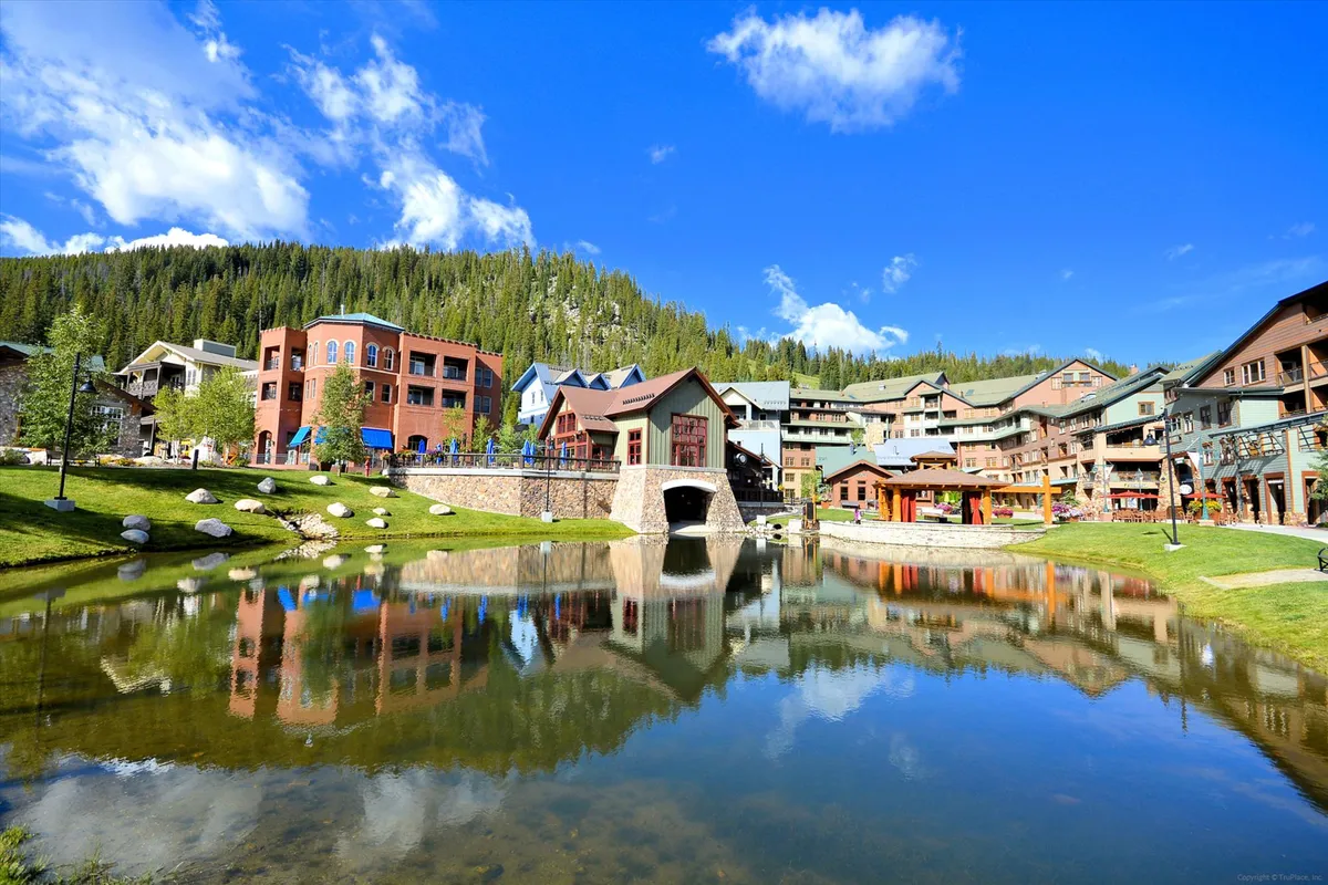 A cheerful exterior image featuring colorful buildings reflected in tranquil water, beautifully landscaped green areas, and inviting walking paths. People are seen enjoying leisurely strolls and picnics, suggesting community vibrancy and easy access to outdoor relaxation beyond the property itself.