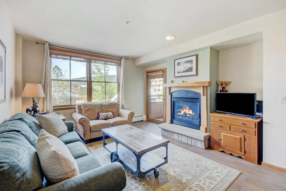 The image showcases a cozy living room interior featuring a warm fireplace, a plush sofa, a stylish coffee table, and expansive windows that frame stunning mountain views. The sunlight is setting outside, casting a golden glow into the room, inviting guests to relax and unwind after a day on the slopes.
