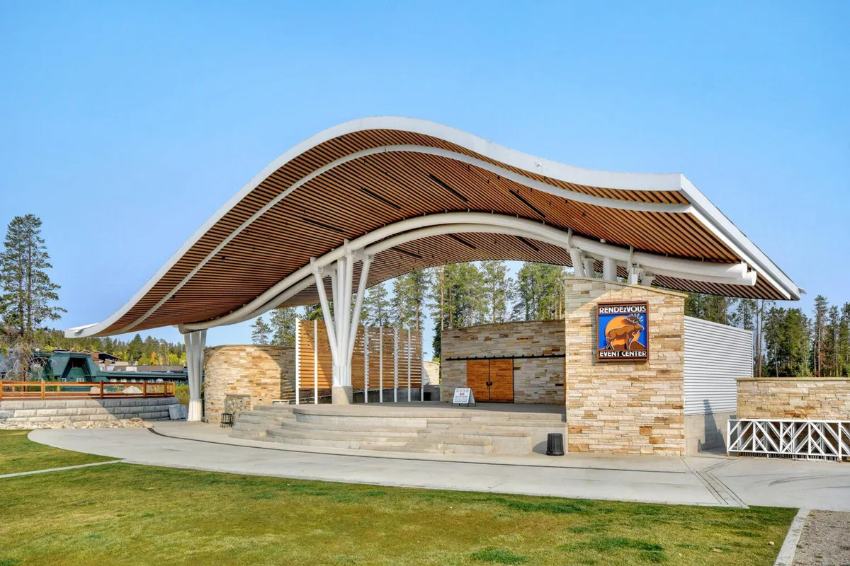 The exterior features a striking curved roof structure, a sturdy stone facade, and warm wooden accents. The area in front of the building appears ready to host outdoor events or welcome guests as they arrive.