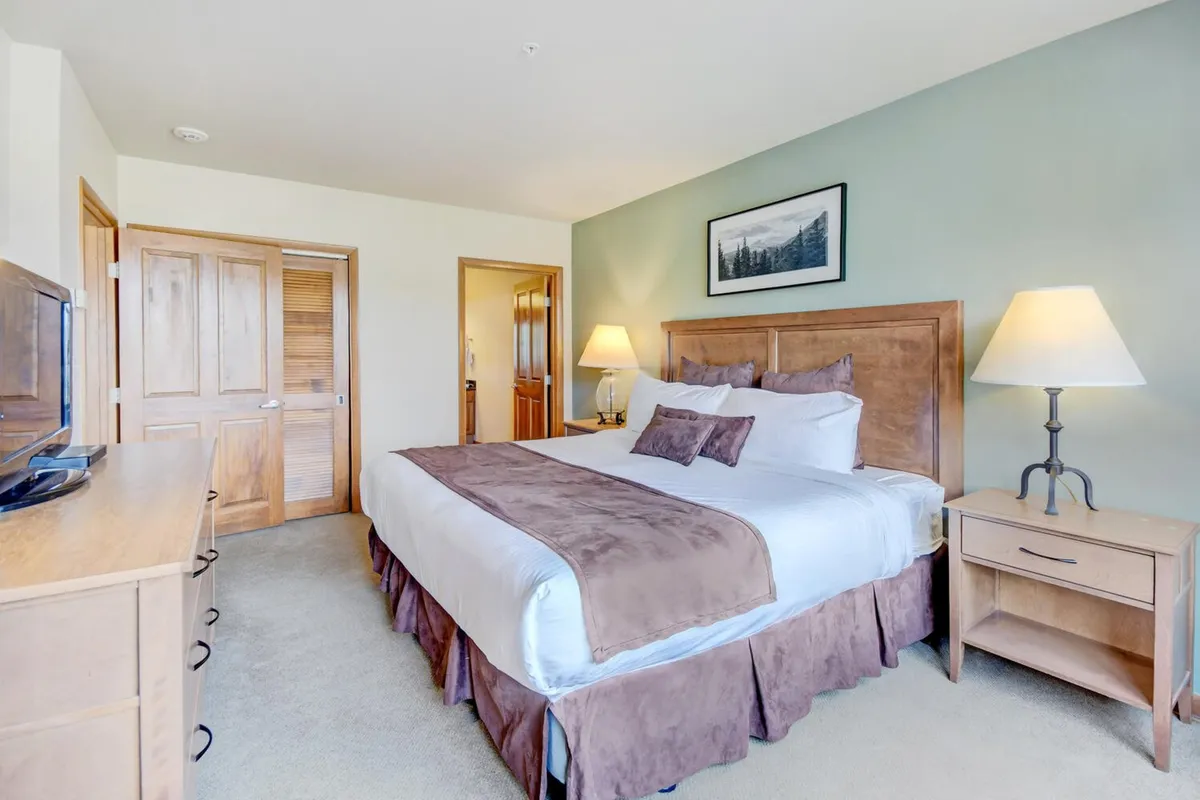 A spacious bedroom with a king-sized bed dressed in soft, inviting linens, complemented by wooden furniture, nightstands, a lamp, and artwork on the wall. The room is filled with natural light, creating a soothing and restful ambiance.