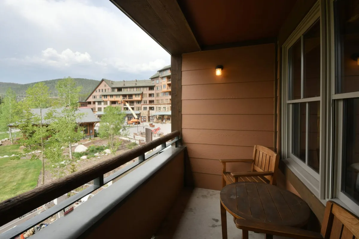 A serene exterior shot featuring a wooden balcony with a sturdy railing, a set of table and chairs, and expansive mountain views in the background. The scene captures the peaceful ambiance of the private covered deck, ideal for morning coffee or evening relaxation while overlooking ski slopes and the lively base village below.