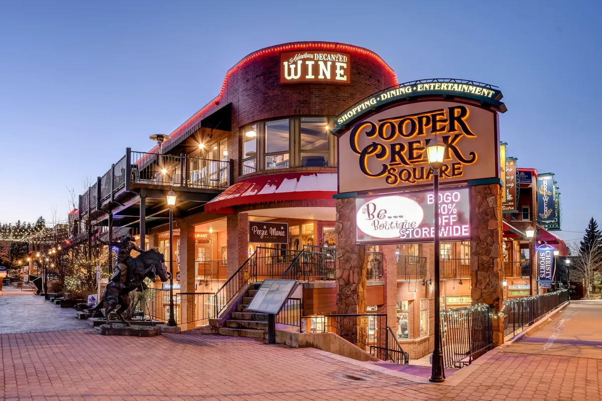 An inviting exterior shot of Cooper Creek Square, showcasing its distinctive signage, a wine shop, a staircase leading to an upper level, decorative lighting, and a contemporary sculpture. The scene is bustling with energy, hinting at a variety of dining, shopping, and entertainment options.