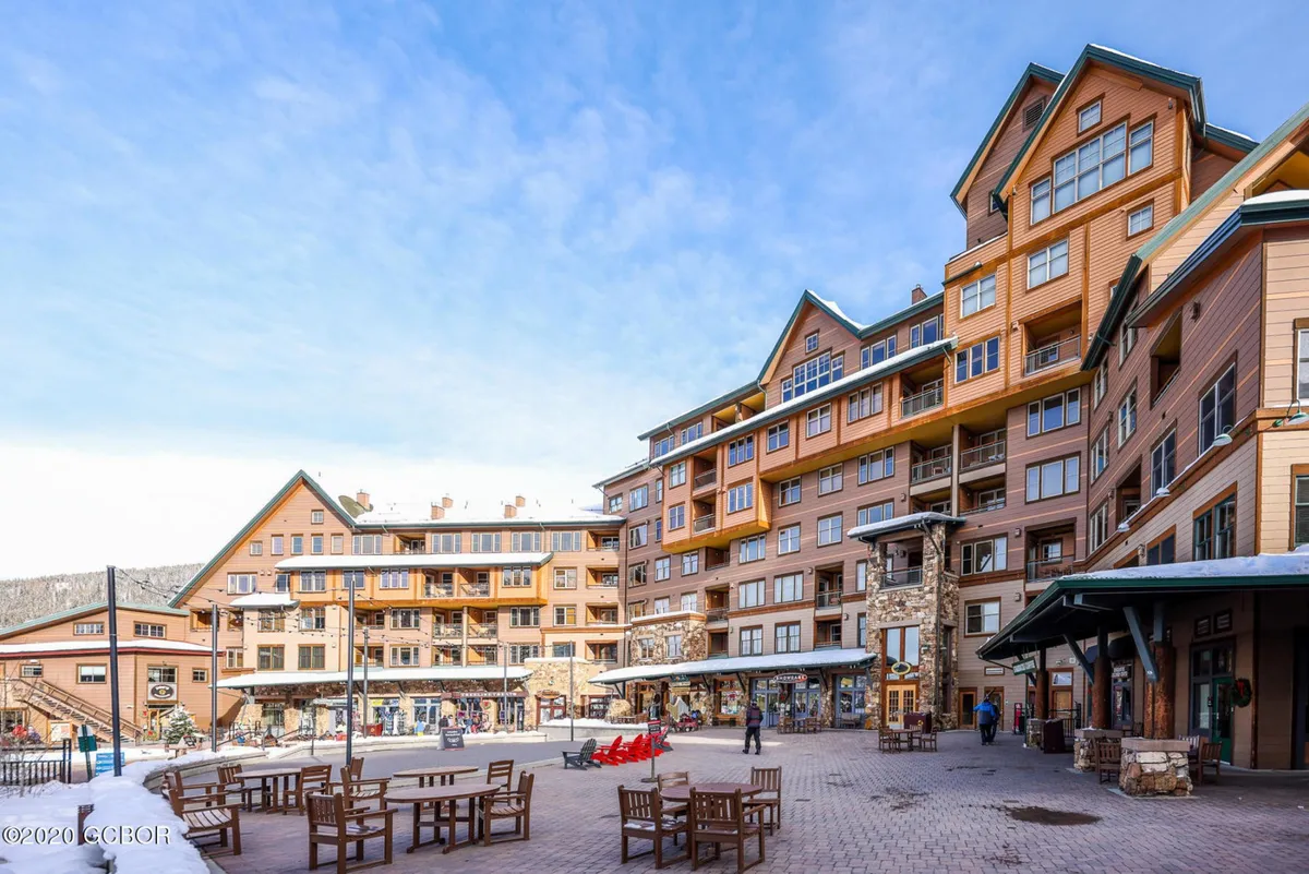 A vibrant exterior shot of the resort's modern architecture, featuring an inviting outdoor seating area bustling with people enjoying the lively mountain village atmosphere. Snow-capped mountains loom in the background, emphasizing the resort's proximity to alpine adventure and village amenities.