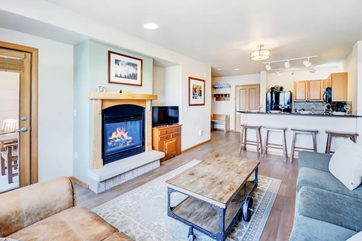 A spacious open-concept living area featuring a plush sofa arranged around a modern gas fireplace, with a coffee table and a view into the connected kitchen area. The scene exudes warmth and comfort, ideal for gathering and relaxing after a day outdoors. The area is well-lit, and the wall-mounted HD TV is visible, providing entertainment options for the entire group.