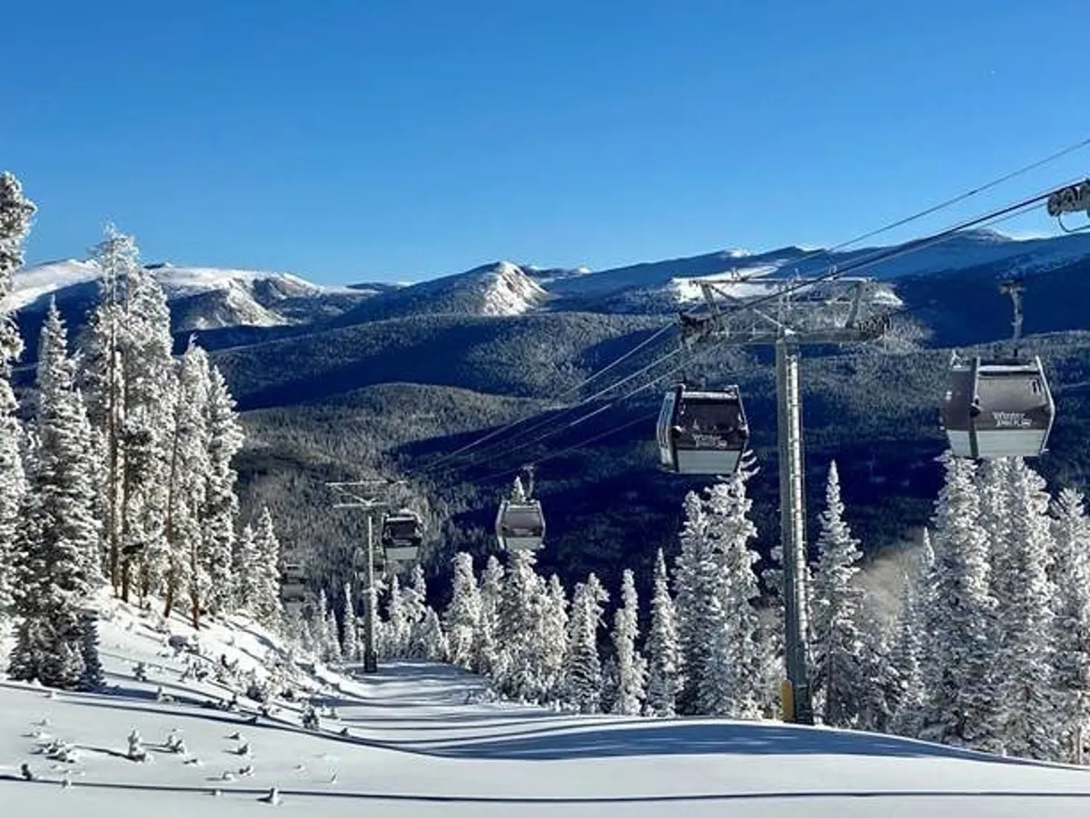 A stunning winter landscape featuring a ski lift ascending over snow-covered trees with a panoramic mountain range in the background. The bright blue sky and pristine snow create an inviting and adventurous winter atmosphere, ideal for skiing and snowboarding.
